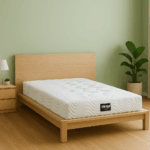 Colchón Teknia Visco sobre cama de madera clara con cabecera lisa, pared verde pastel, mesilla con lámpara y planta decorativa