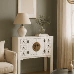Consola blanca con puertas y cajones dorados colocada en sala moderna, con pared verde suave, sofá beige y lámpara de mimbre.