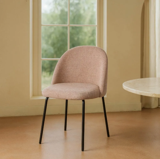 Silla tapizada color rosa con patas metálicas negras junto a mesa redonda y ventana