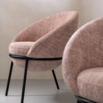 Sillón moderno tapizado en tela con reposapiés a juego y patas metálicas negras, disponible en gris, beige y rosa pastel.